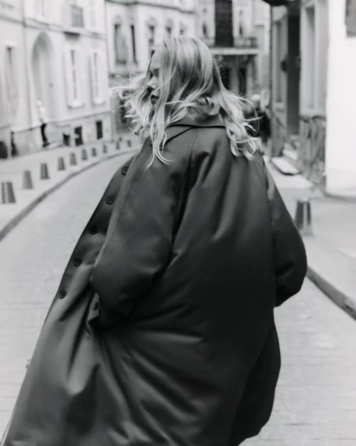 Black and white shot of a woman with long hair walking on a city street wearing an oversized coat in an urban setting.