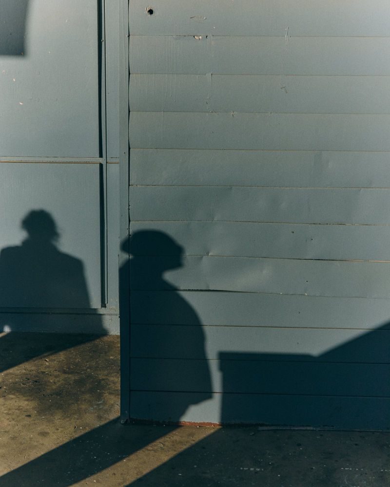 Two silhouetted figures cast shadows on a weathered grey building exterior with horizontal siding and a nearby concrete surface.