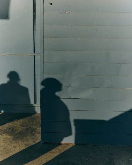 Two silhouetted figures cast shadows on a weathered grey building exterior with horizontal siding and a nearby concrete surface.