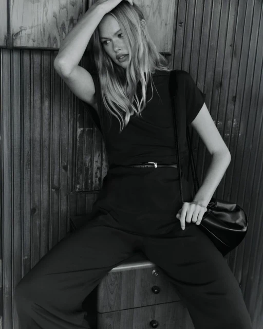 Black and white portrait of a woman in a wrap top and pants sitting on a dresser in front of a wooden wall.