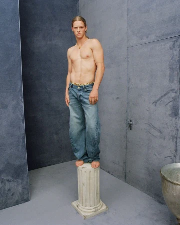 Shirtless male model with blond hair poses on a pillar in a gray indoor setting, wearing blue jeans.
