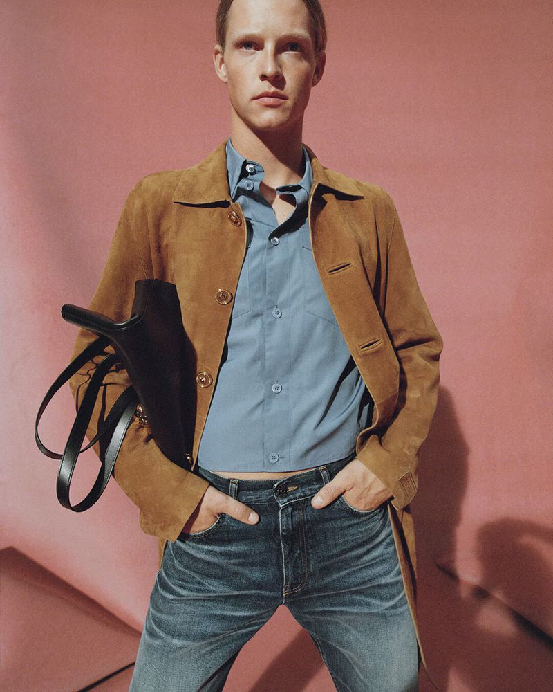 Man in blue button-down shirt, brown suede jacket, and jeans poses indoors against a pink paper background, carrying a black handbag.