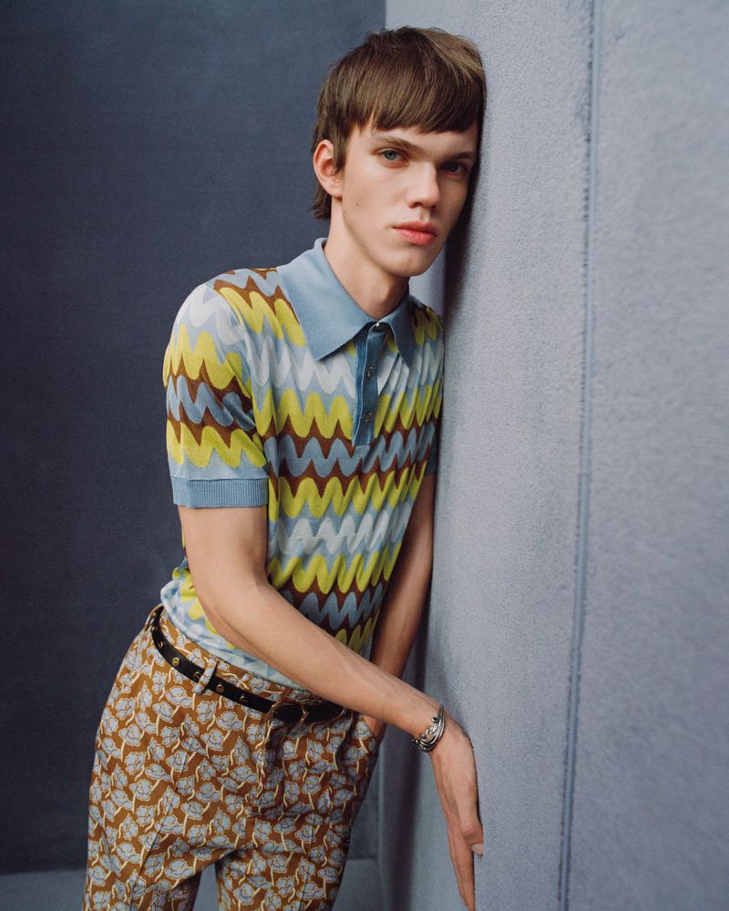 Stylish young man in patterned polo shirt and trousers leans against a blue wall in a studio setting, fashion portrait.