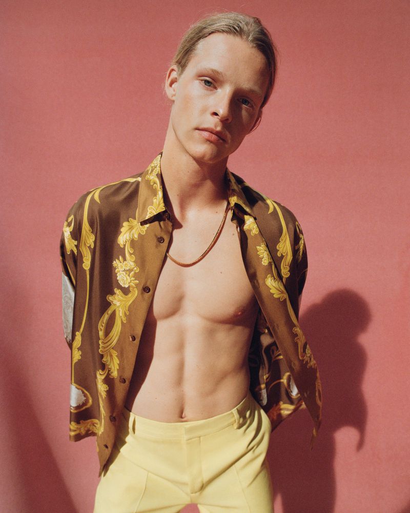 An adult male model poses in an open brown and gold patterned shirt with yellow pants against a pink studio backdrop, highlighting fashion and style.