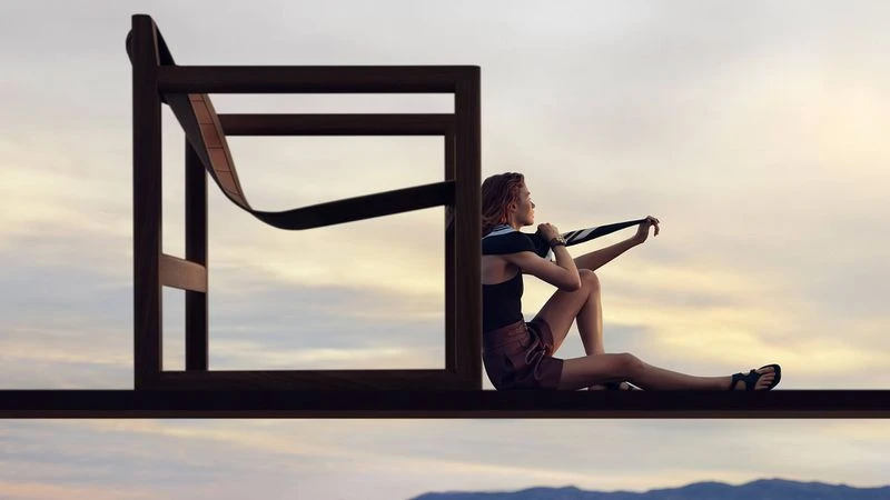 A woman sits on a brown I-beam construction form in front of a deconstructed wooden chair against a cloudscape backdrop at sunset.