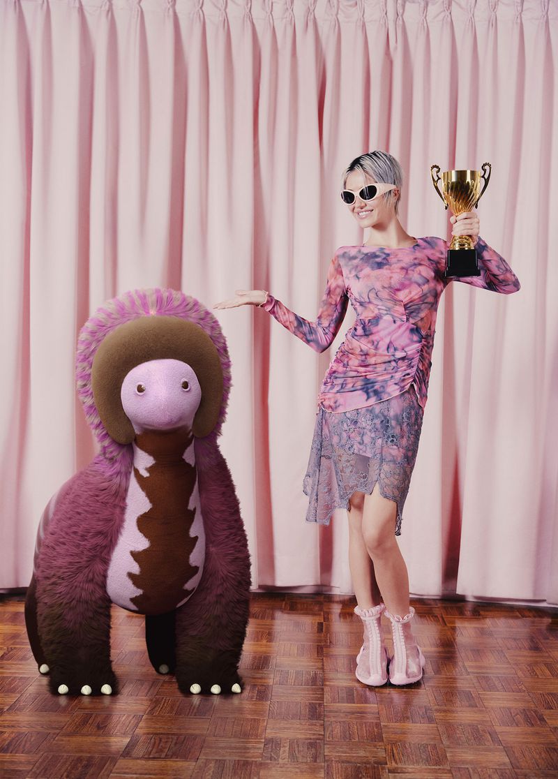 Woman in floral dress and sunglasses poses with a stuffed dinosaur toy and a gold trophy against a pink curtain backdrop.
