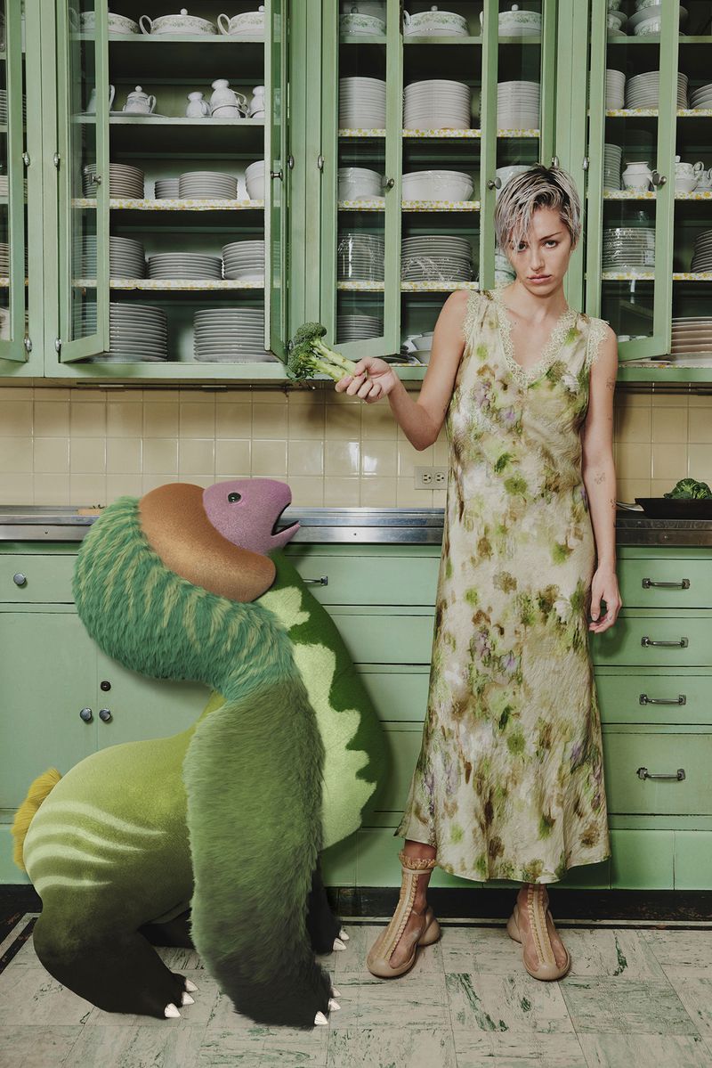 Woman in floral dress holds broccoli next to a surreal green monster in a vintage mint-green kitchen with white dishware displayed.