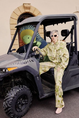 An adult female model in a green camouflage outfit poses next to a black utility vehicle with a mascot in the passenger seat outdoors.
