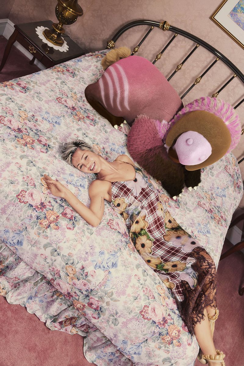 Woman in patchwork dress reclines on a floral bed with novelty pillows. A nightstand with a lamp and framed art are in the scene.
