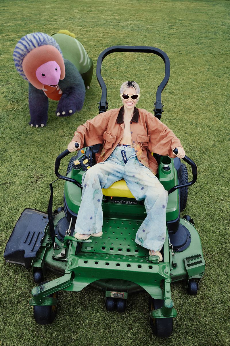 Adult female poses on green lawn mower next to plush monster toy on manicured lawn, wearing vintage sunglasses, jacket, and bell-bottom jeans.