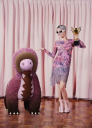 Woman in pink mesh patterned dress and sunglasses holding a gold trophy next to a fuzzy dinosaur creature before pink curtains.