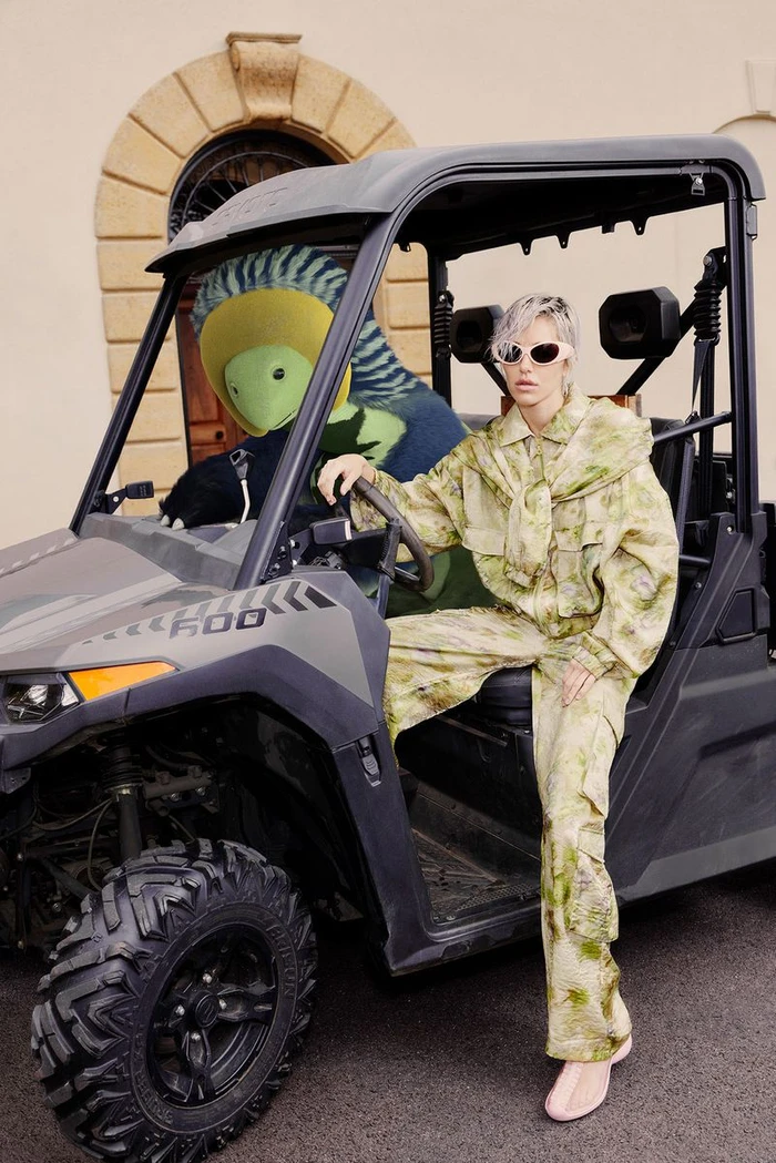 Model wearing patterned green outfit sitting in a grey utility terrain vehicle next to a large plush toy