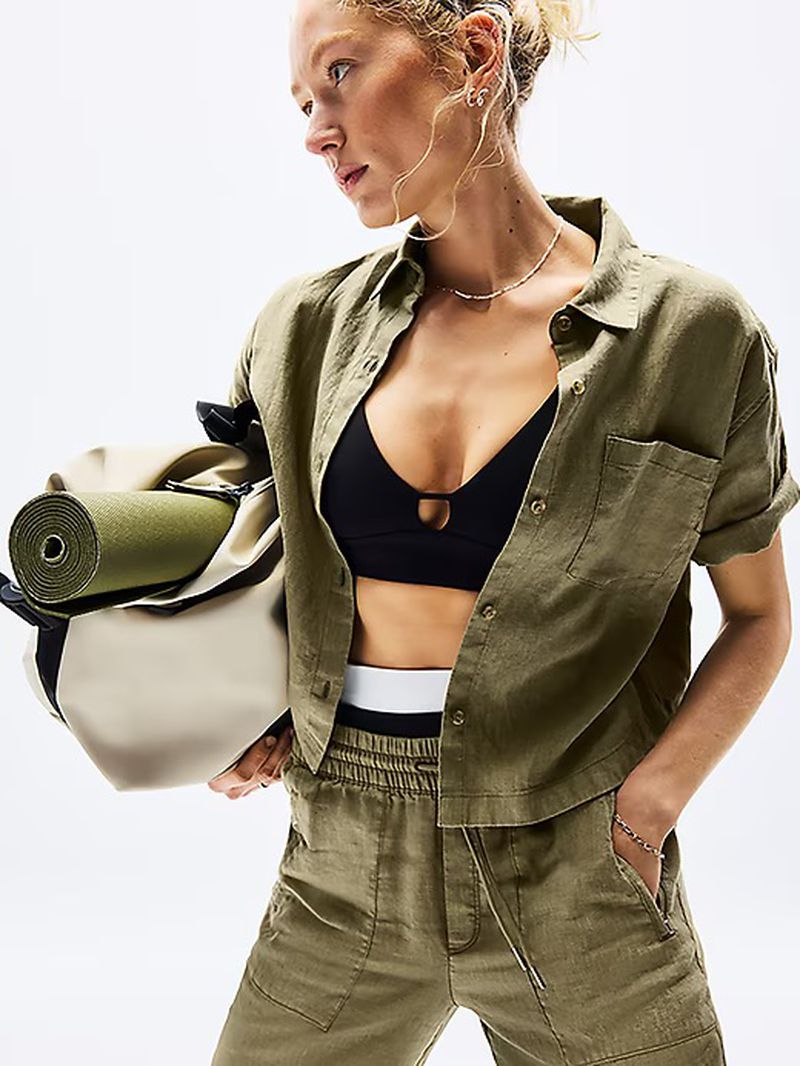 Woman in olive button-down shirt and black sports bra carrying a yoga mat in a beige tote bag against a bright white background.