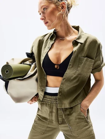 Woman in olive button-down shirt and black sports bra carrying a yoga mat in a beige tote bag against a bright white background.