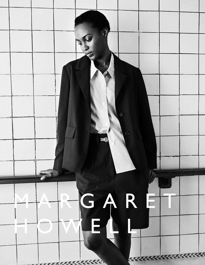 An adult woman in a black blazer and shorts stands against a white tiled wall with rail in a fashion editorial shot in black and white.