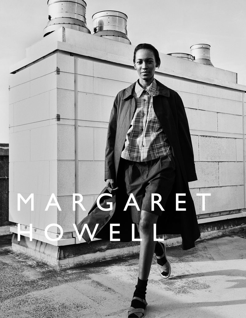 Black and white fashion shot of a woman in a plaid shirt, shorts, sandals, and trench coat standing on a rooftop with vents.