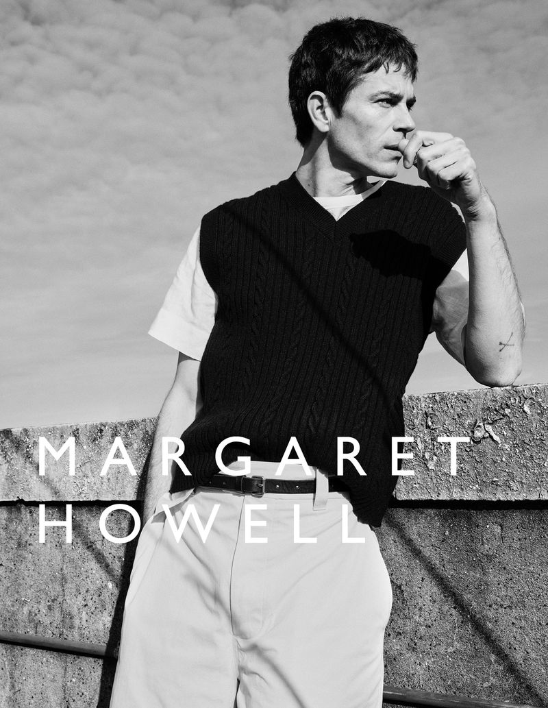 A man poses outdoors in a black vest and white shirt, leaning against a concrete barrier. The text 'MARGARET HOWELL' is visible.