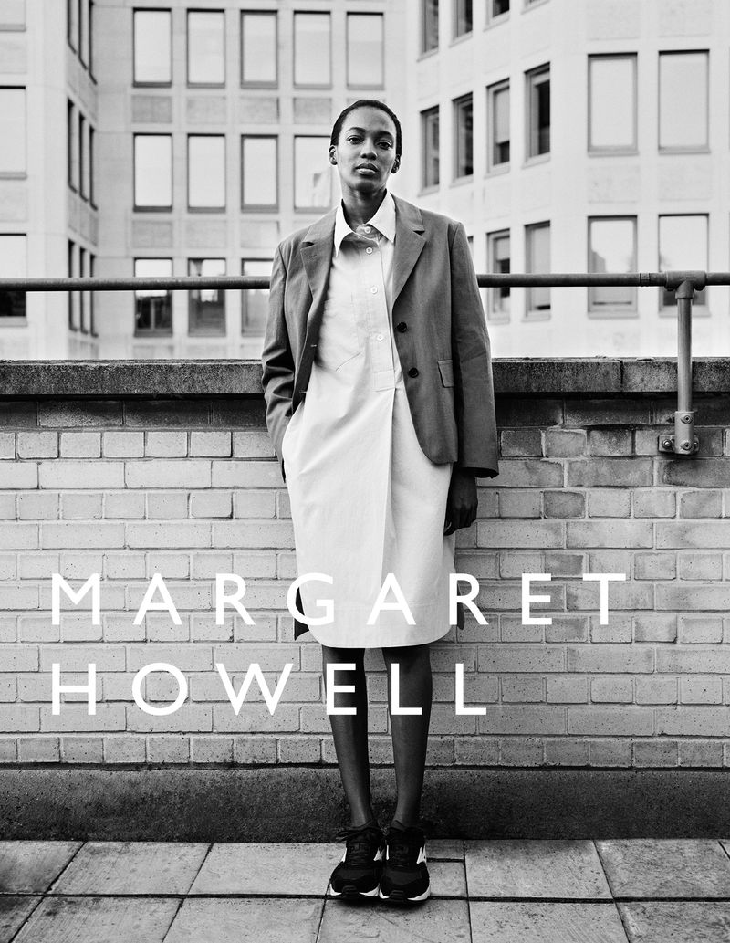 Monochromatic portrait of a black female model in a blazer and dress, standing in front of a brick wall with the MARGARET HOWELL logo.