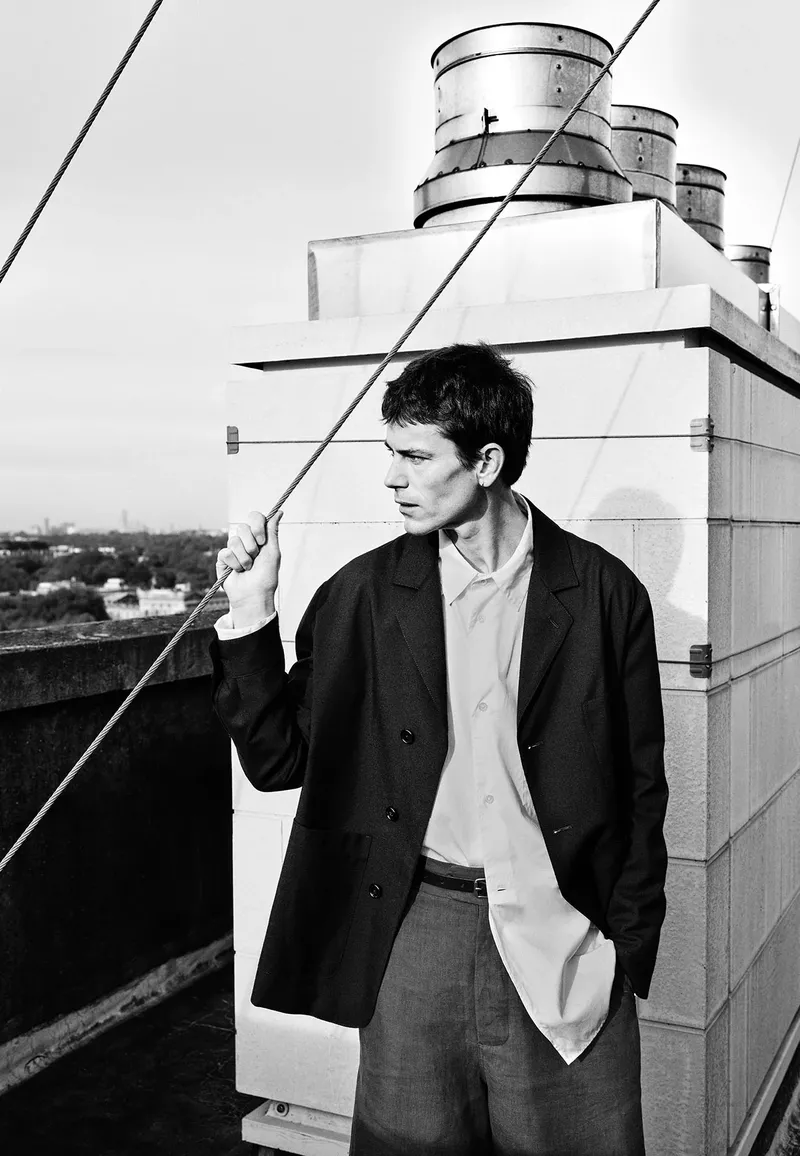 Monochrome portrait of a stylish adult male in a blazer and collared shirt, standing on a rooftop with city buildings visible in the distance.