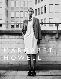 Model standing against a brick wall and building exterior wearing a blazer and dress for Margaret Howell S/S 2025 Campaign