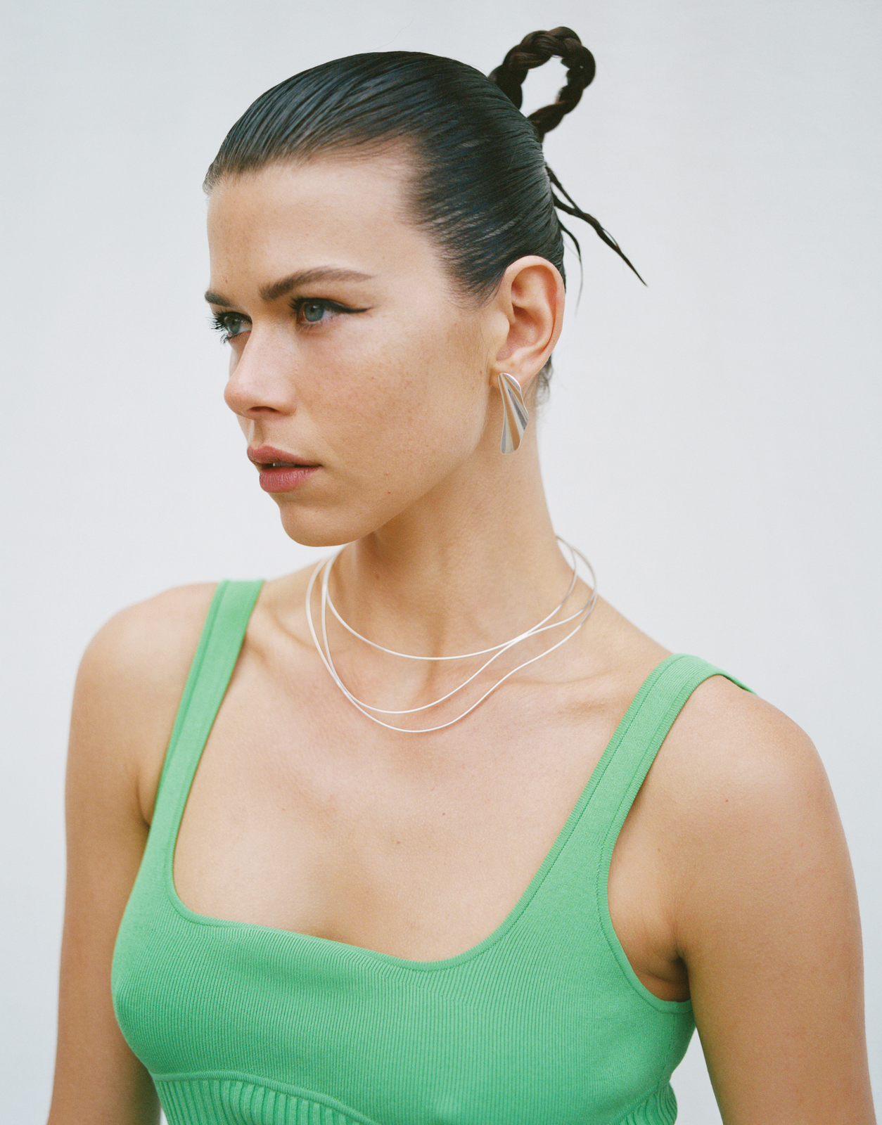 Close-up of a woman with her hair in a twisted bun, wearing a green top and silver jewellery, against a plain background.