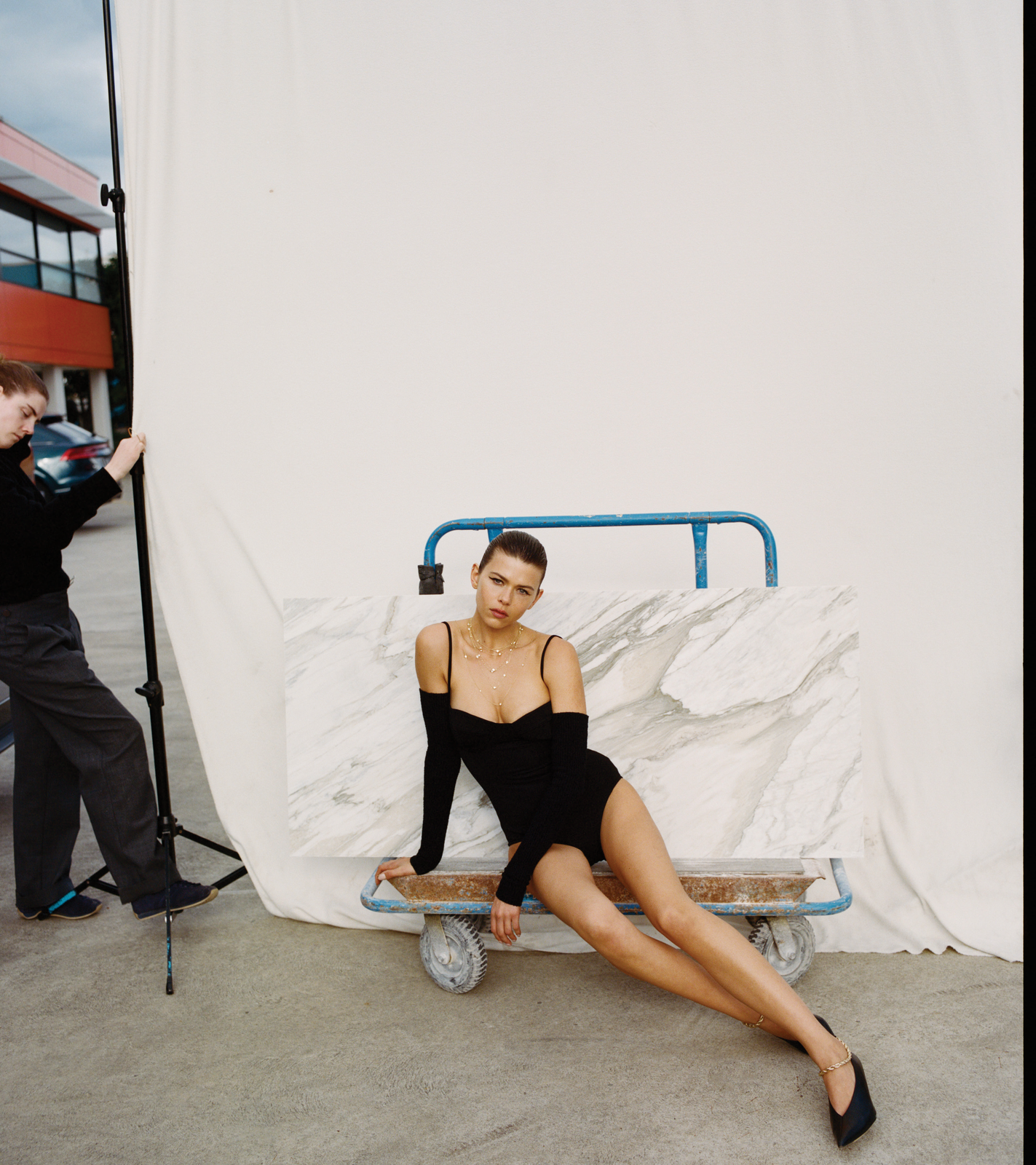 Woman in black bodysuit poses on dolly with marble slab near assistant holding backdrop during outdoor fashion photoshoot.