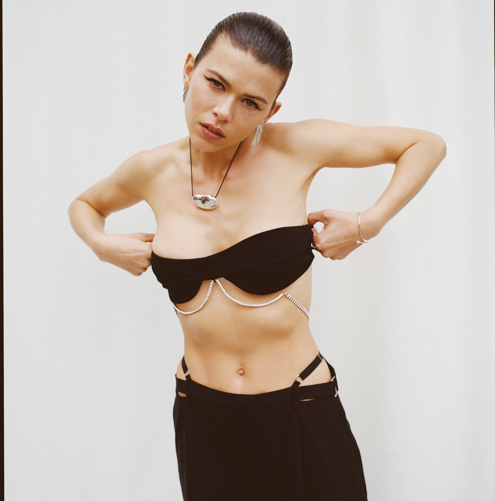 Fashion shot of a young woman in a bra-like top and low-slung skirt against a white background, accessorized with a necklace and bracelets.