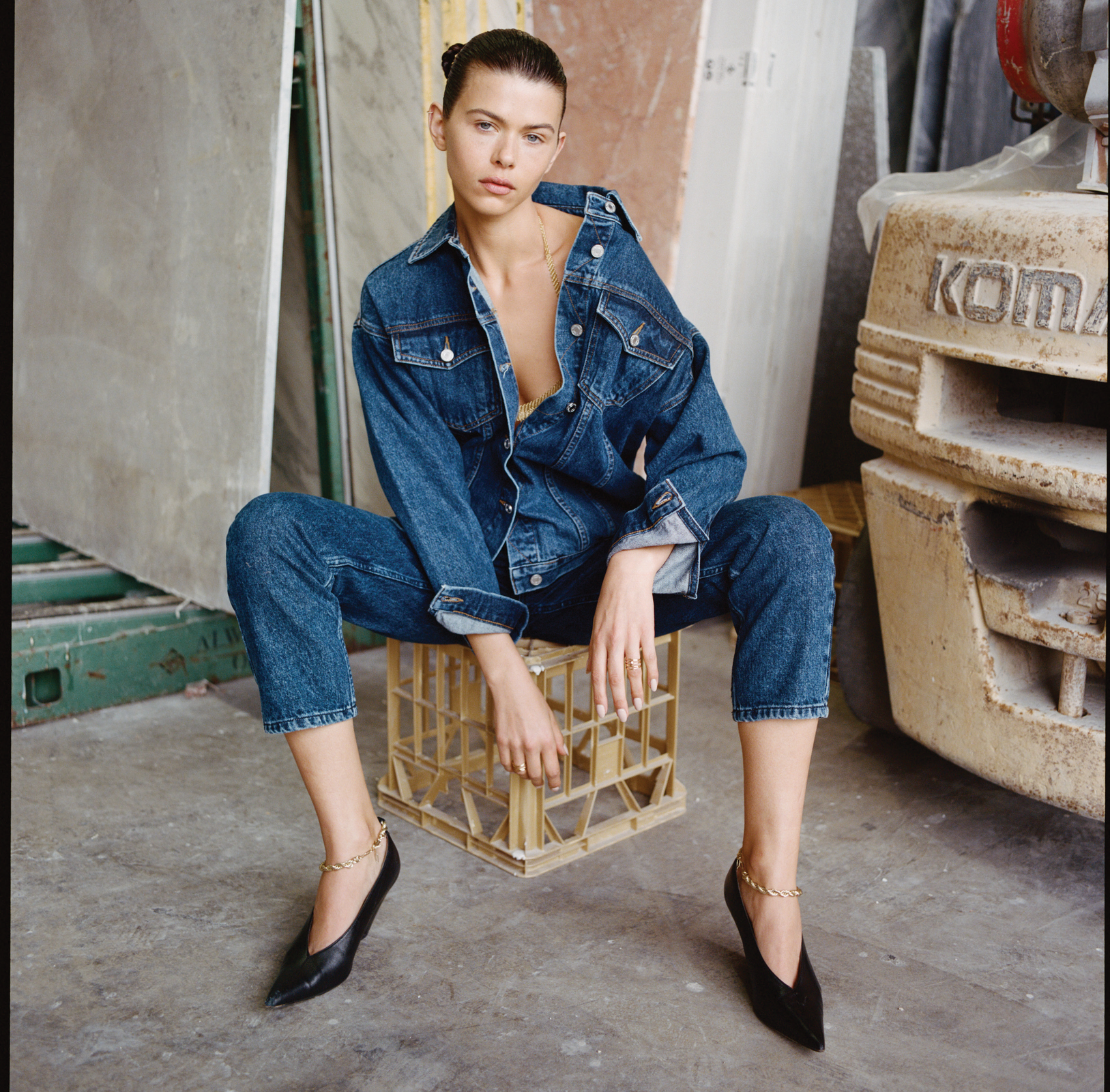A woman in a denim jacket and jeans sits on a plastic crate, showcasing a casual yet stylish look in an industrial environment.