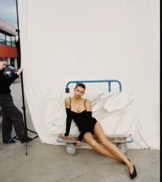 Georgia Fowler seated on a blue utility cart wearing a black bodysuit against a white backdrop outdoors