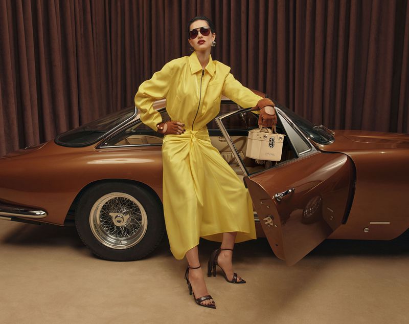 A stylish woman in a yellow dress poses beside a classic brown car with a cream-colored handbag and curtain backdrop in studio lighting.