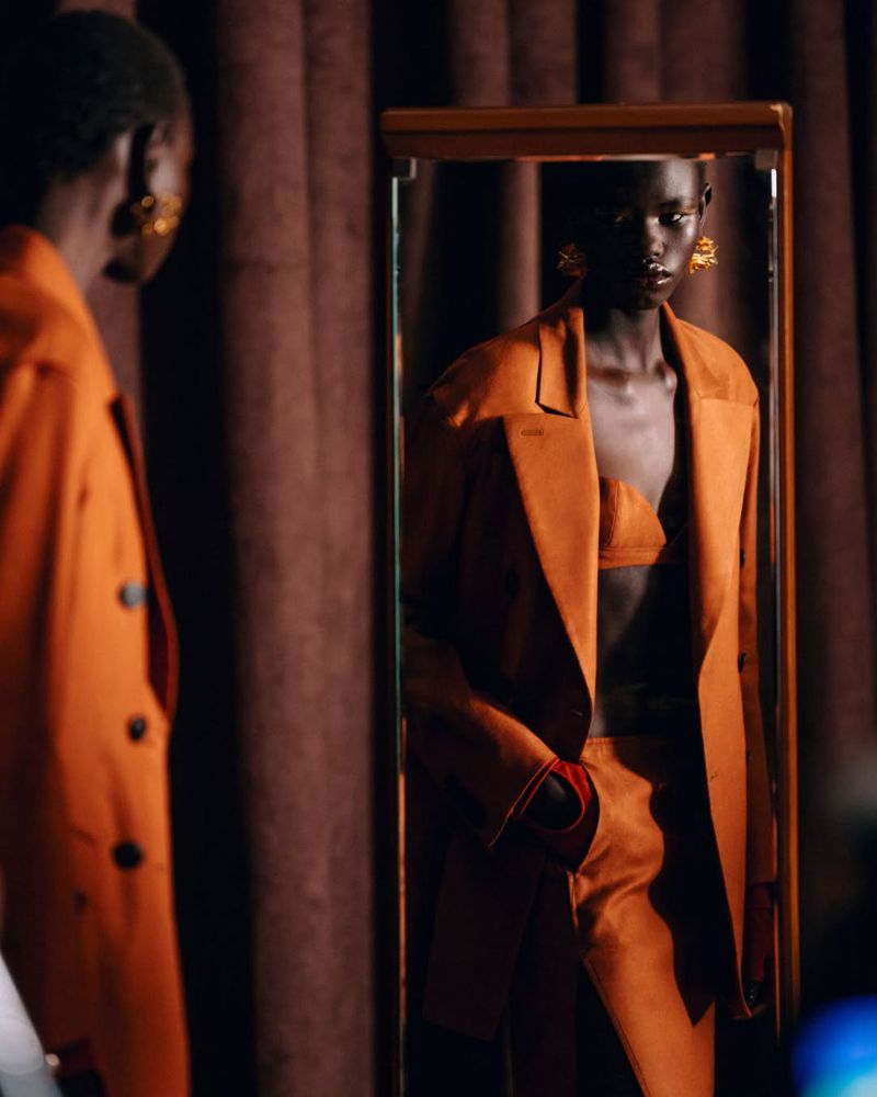 Elegant African woman in an orange suit reflected in a mirror, showcasing fashion and style in an indoor setting with brown curtains.