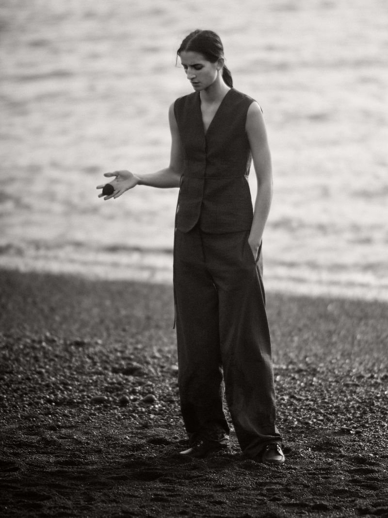 Woman stands on a dark sand beach in tailored vest and wide leg pants, holding a dark round object, with a water background.