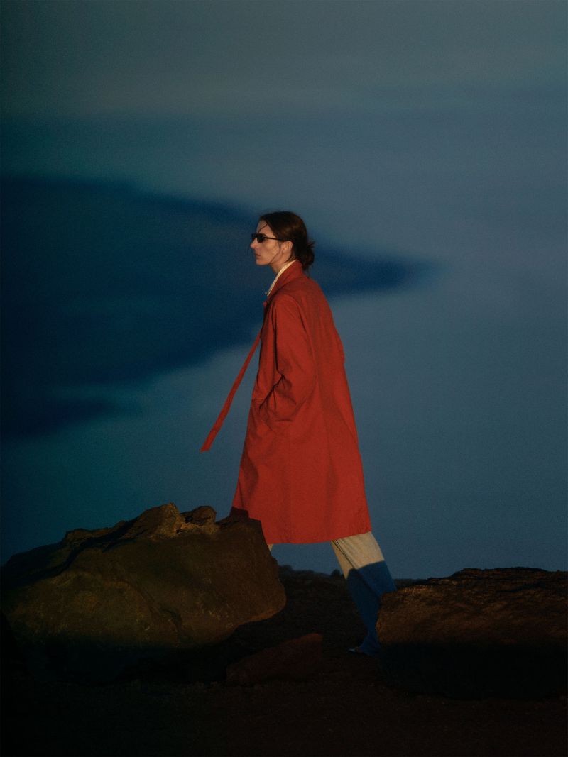 A woman in a red coat walks on rocks against an abstract blue background, photographed at eye-level in a fashion editorial style.