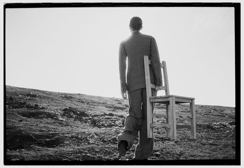 Monochrome image of a person in a suit carrying a wooden chair across a sparse landscape under a bright sky, captured in a wide shot.
