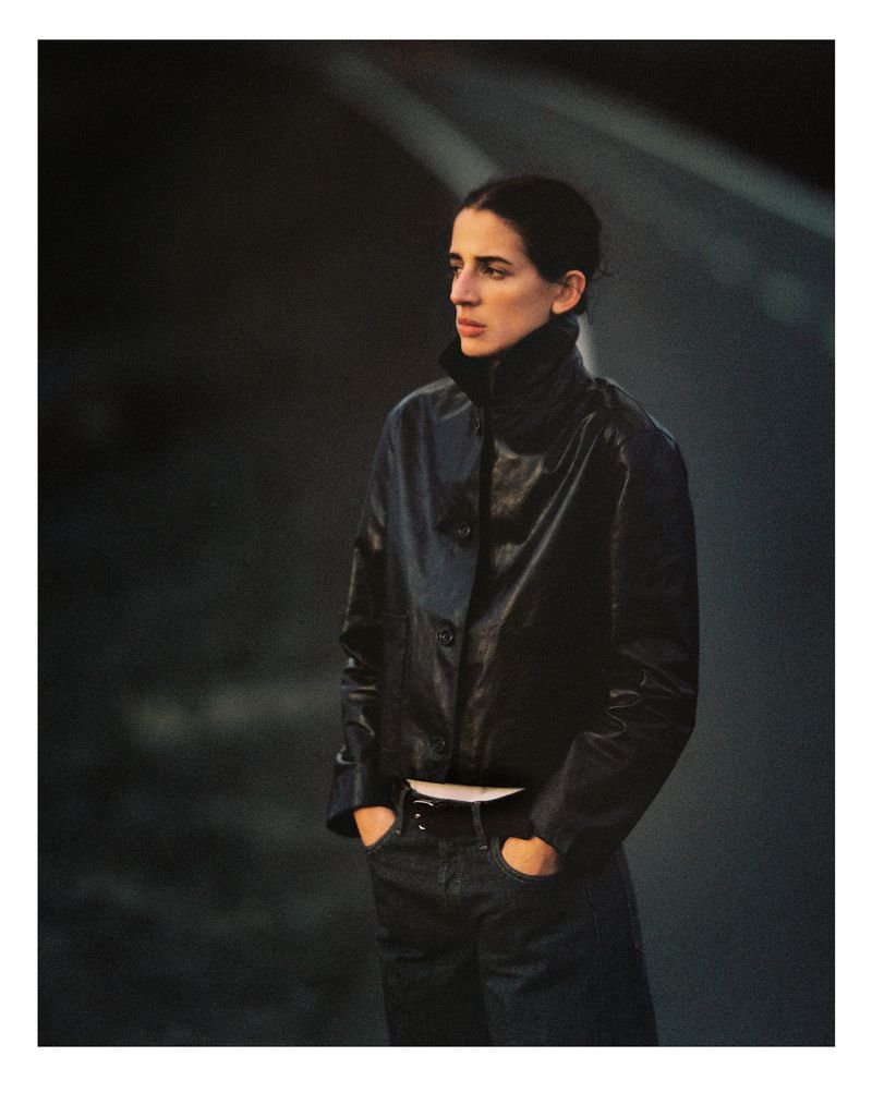 Portrait of a woman in a black leather jacket and jeans standing outdoors with hands in pockets against a dark, abstract background.