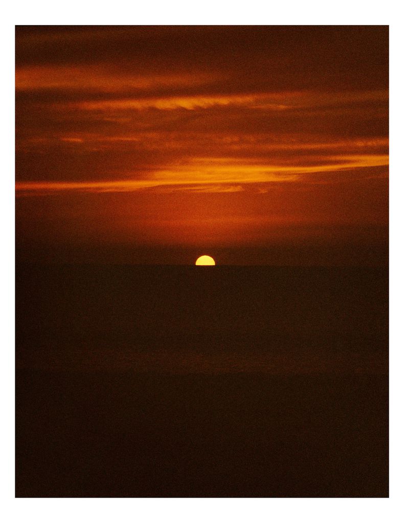 Dramatic sunset over a dark ocean, with a half-visible sun and colorful orange, yellow and brown clouds filling the sky above.