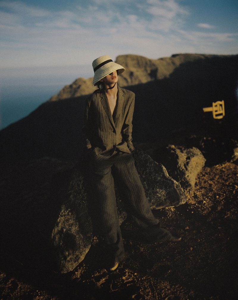 Model wearing a straw hat and tailored trousers seated on rocks against mountainous terrain at dusk