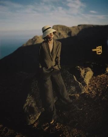 Model wearing a straw hat and tailored trousers seated on rocks against mountainous terrain at dusk