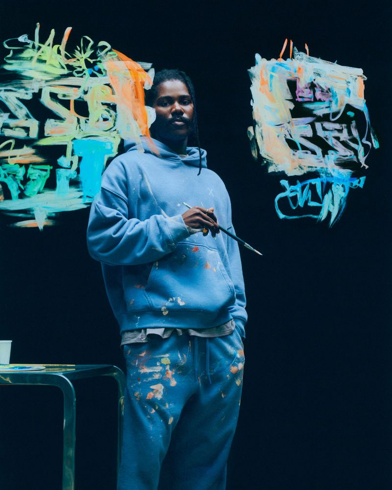 Man in light blue hoodie and sweats holding a paintbrush in front of abstract graffiti art on a dark background in a studio setting.