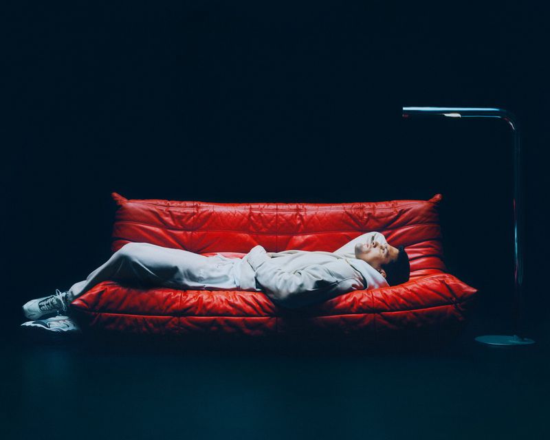 An adult male wearing a white sweatsuit lies on a red leather couch with a silver arc floor lamp in a minimalist studio setting.