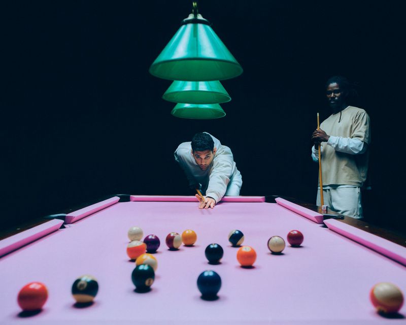 Two men, one aiming with a cue stick on a pink pool table with scattered balls, beneath green lighting in a dark room.