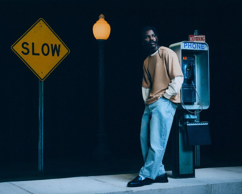 Man leans against a vintage phone booth at night with a 'SLOW' street sign, illuminated by a glowing street lamp in an urban setting.