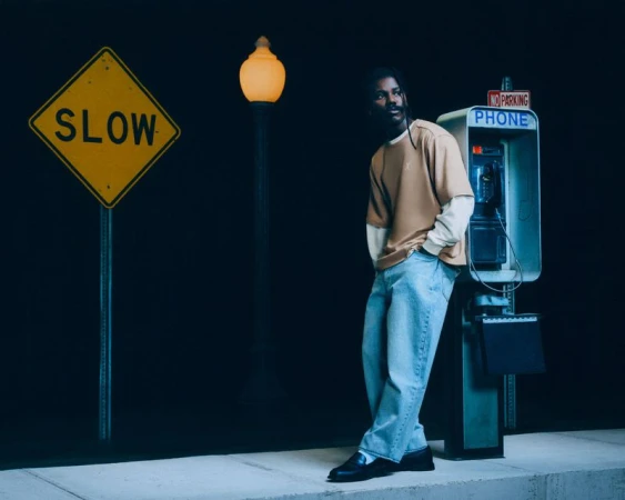 Man leans against a vintage phone booth at night with a 'SLOW' street sign, illuminated by a glowing street lamp in an urban setting.
