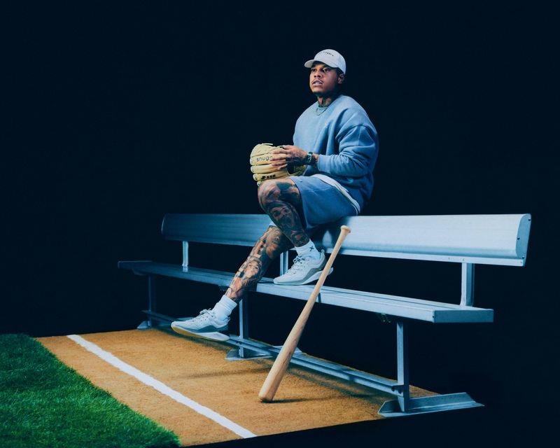 Tattooed man in sweatshirt and baseball cap sits on a bench with baseball glove and bat in an indoor studio setting with dark background.
