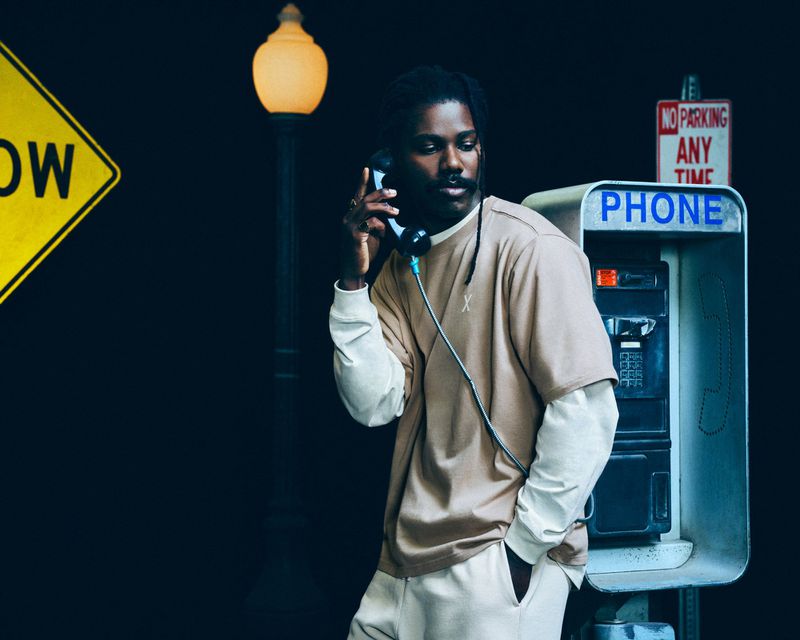 Man in a beige T-shirt uses a vintage payphone in a dimly lit urban setting with a yellow street sign and lamppost.