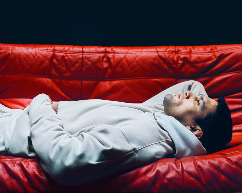 A man in a white sweatshirt is reclining on a red leather couch against a dark background, looking to his right side.
