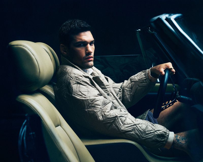 Man driving a convertible in a low-lit setting, wearing a patterned jacket, showcasing modern automotive lifestyle and fashion.