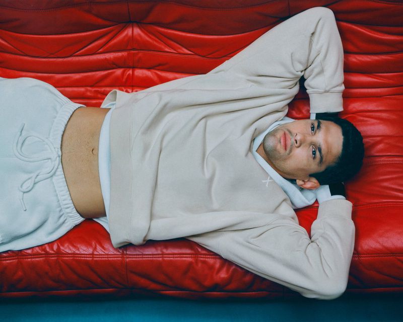 Young adult male lying on a bright red leather couch wearing a beige hooded sweatshirt and sweatpants in a horizontal portrait.