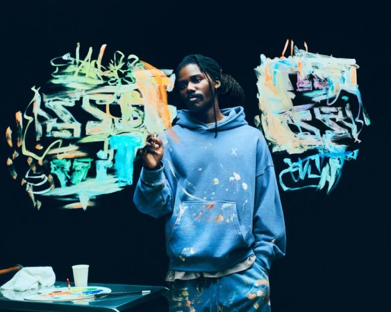 Portrait of a Black male artist with dreadlocks in a paint-splattered hoodie, holding a paintbrush in front of colorful abstract art.