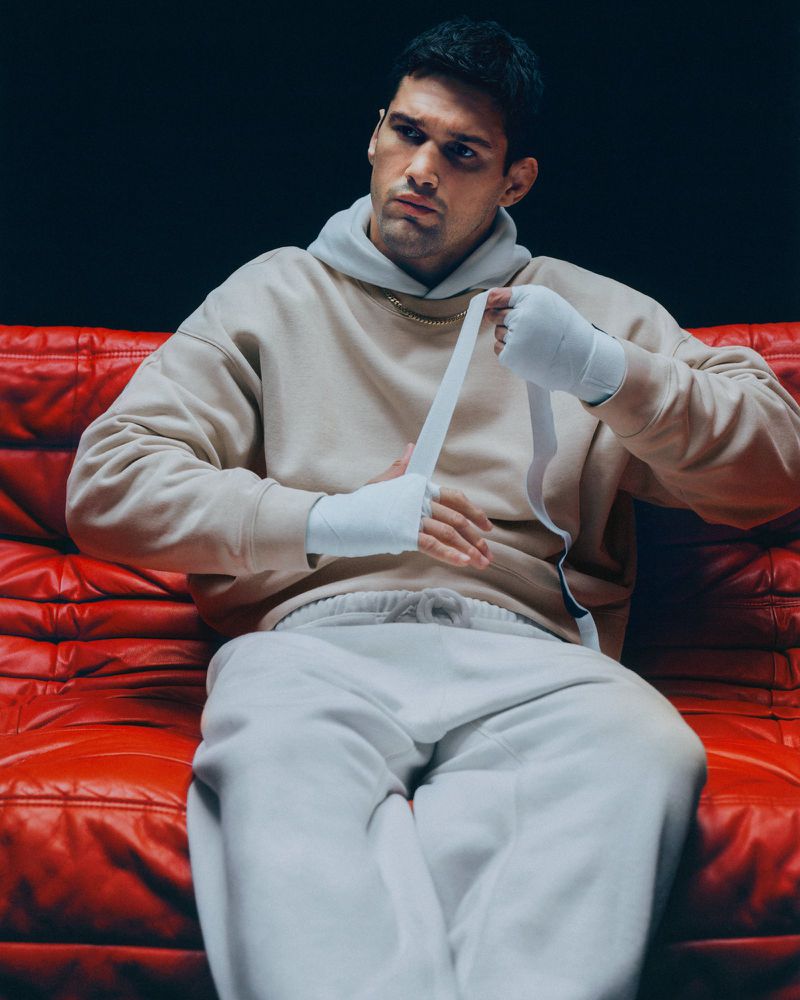 Man wraps his hands on a red leather couch in photo studio. He wears a beige hoodie and sweatpants and has a neutral expression.